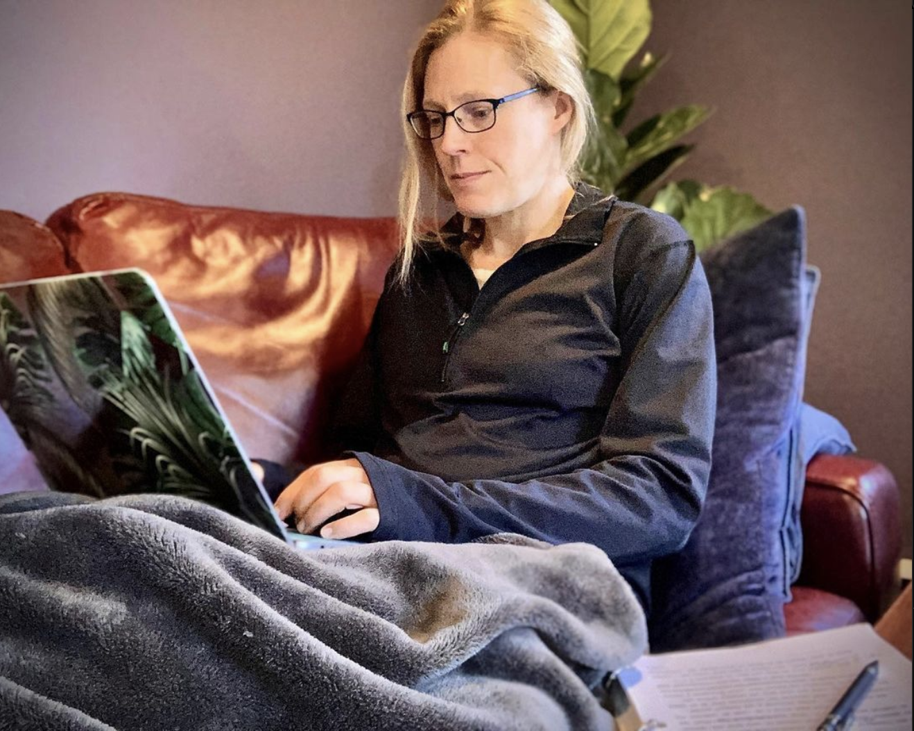 Photo of the author sitting on a couch, looking at the screen of the macbook sitting in her lap. She's wearing glasses.